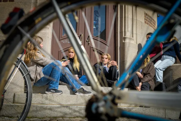 Students sitting on a staircase outside a gate with a bicycle wheel in front of them.
