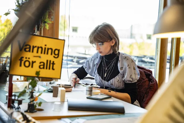 Student working at a desk. In the foreground is a yellow sign with black text: Warning for everything.